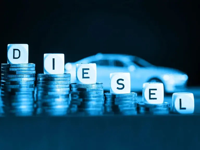Mehrere Stapel Geldmünzen, die von links nach rechts niedriger werden. Auf den Münzen sind Würfel platziert, die das Wort Diesel ergeben. Im Hintergrund ein Auto