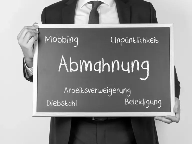 Ein Mann, der eine Tafel vor sich hält auf der steht: Abmahnung in der Mitte und weitere Worte: Mobbing, Unpünktlichkeit, Arbeitsverweigerung, Diebstahl, Beleidigung