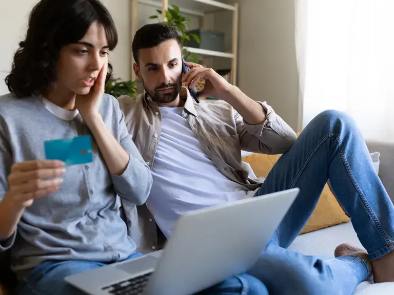 Ein Paar das auf ein Laptop blickt. Die Frau hält eine Kreditkarte in der Hand, der Mann telefoniert