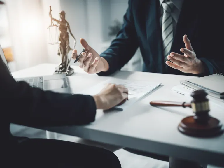 Ein Schreibtisch an dem 2 Menschen sitzen und beraten. Auf dem Schreibtisch juristische Utensilien wie die Justitia-Statue und ein Richterhammer