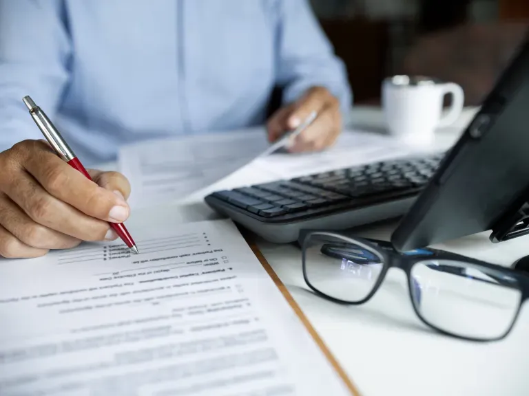 Ein Schreibtisch auf dem ein Laptop steht, eine Brille im Vordergrund. Im Hintergrund ein Mann der mit einem Kugelschreiber auf einem Papier etwas schreibt.