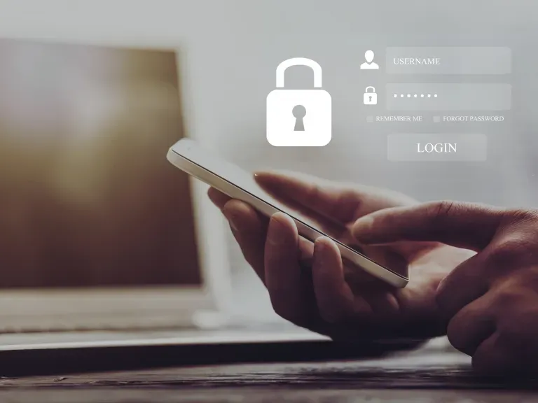 Im Hintergrund ein Laptop. Im Vordergrund Hände die ein Handy bedienen. Über den Handy schweben digitale Eingabefelder von Login Daten