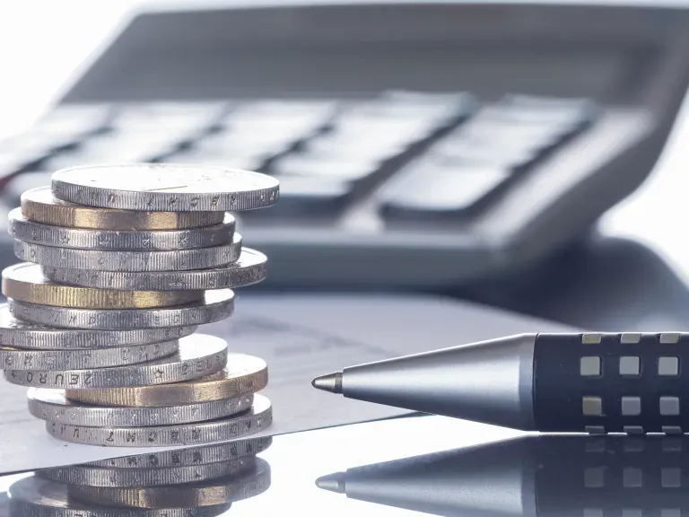 Im Vordergrund ein Stapel Münzen und die Spitze eines Kugelschreibers. Im Hintergrund ein Taschenrechner