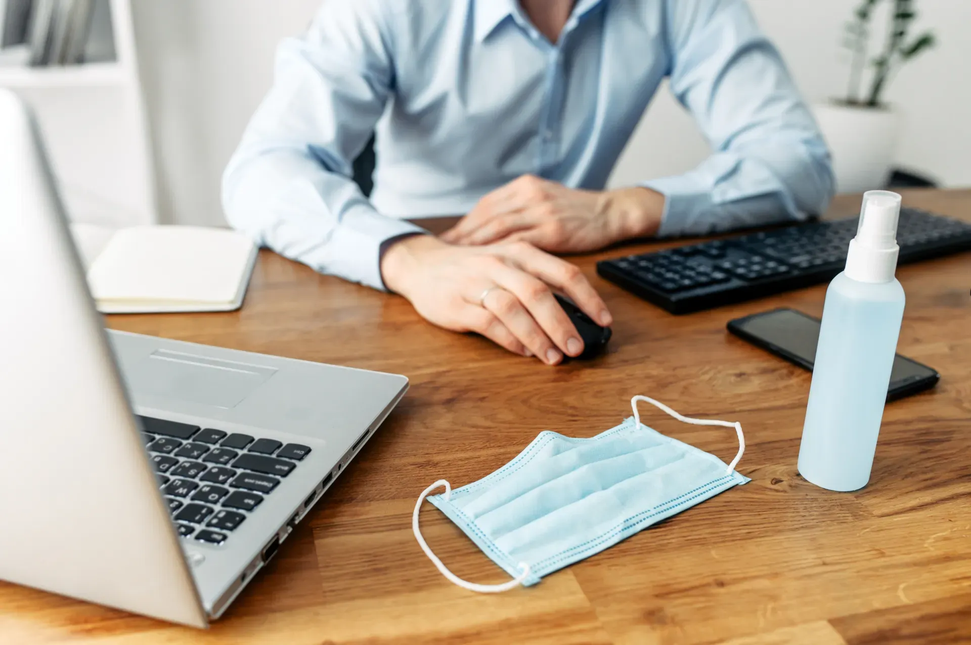 Ein Schreibtisch auf dem ein Laptop, eine Maske und Desinfektionsspray stehen. Hinter dem Schreibtisch ein Mann, der eine Maus bedient.